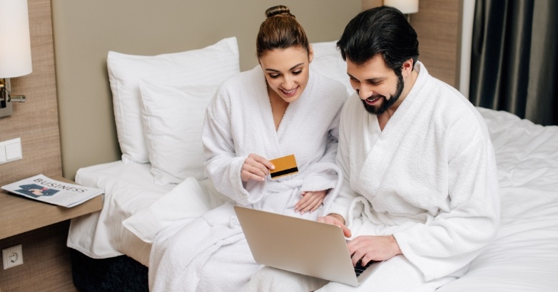 A man and woman in bathrobes are seated on a bed, working on a laptop related to hotel lead generation strategies.