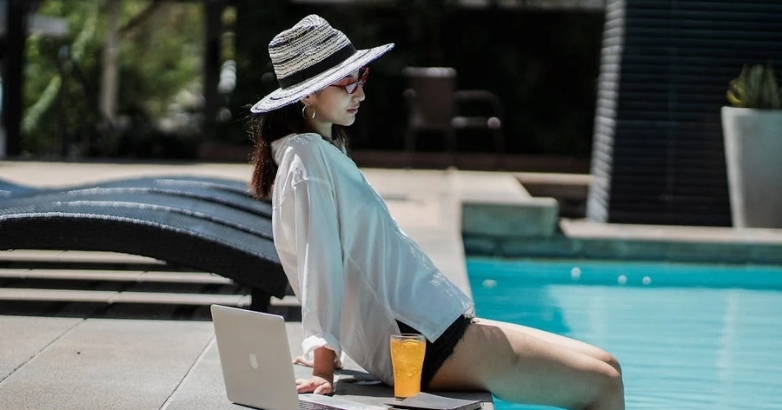 A woman in a hat sits by a pool, working on a laptop, promoting hotel branding with local influencers.