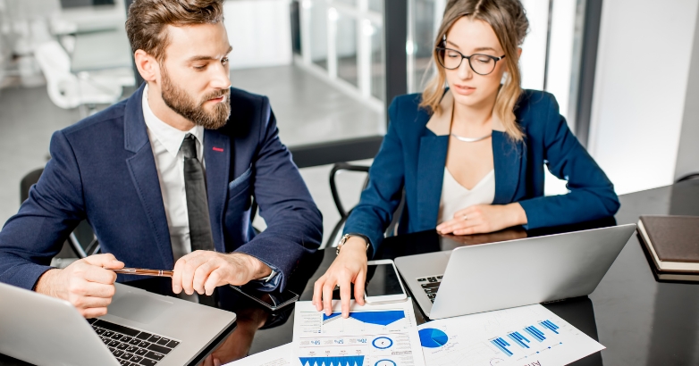 Two business professionals seated at a table, working on laptops during a hotel marketing audit competitive review meeting.