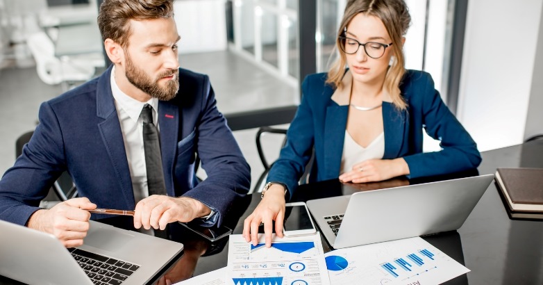 Two business professionals seated at a table, working on laptops during a hotel marketing audit competitive review meeting.