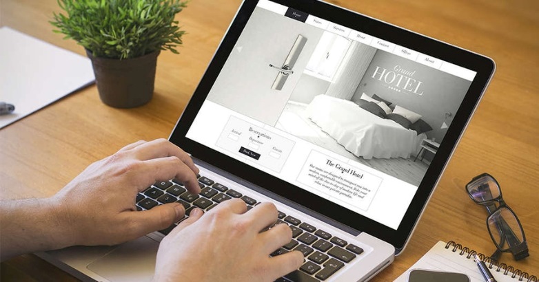 A person uses a laptop to view a hotel booking website at a wooden desk with a plant, notebook, and pen nearby, exploring options influenced by the Hotel Digital Marketing Cycle and effective Search Engine Optimization.