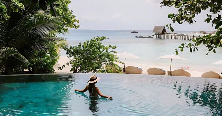 Woman in a straw hat in an infinity pool overlooking a tropical beach and pier.