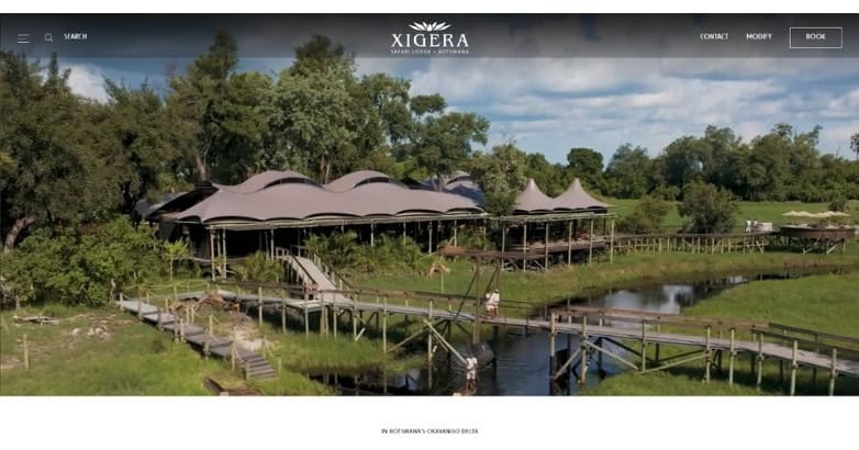 Aerial view of a modern lodge with a unique roof design, surrounded by lush greenery and a winding pathway over water.