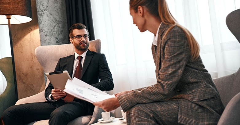 Two professionals in business attire sitting in chairs, engaged in a conversation about hotel marketing collaboration.