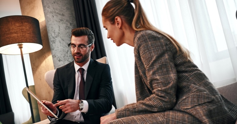 A man and woman in professional clothing sitting on a couch, engaged in a conversation about hotel website design.