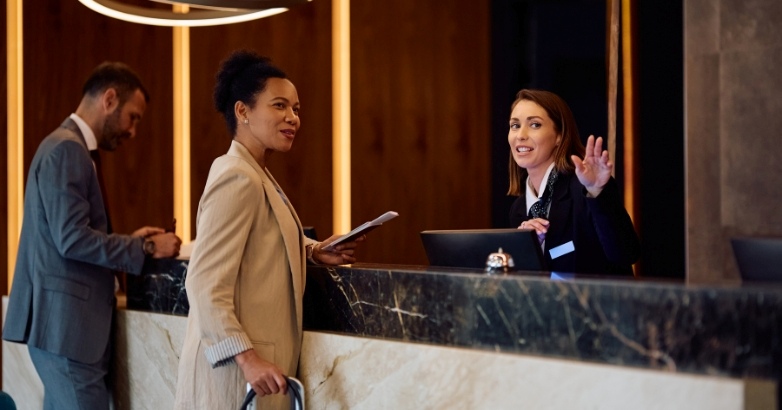A business-suited woman stands at the front desk, focusing on hotel marketing KPIs and customer acquisition costs.