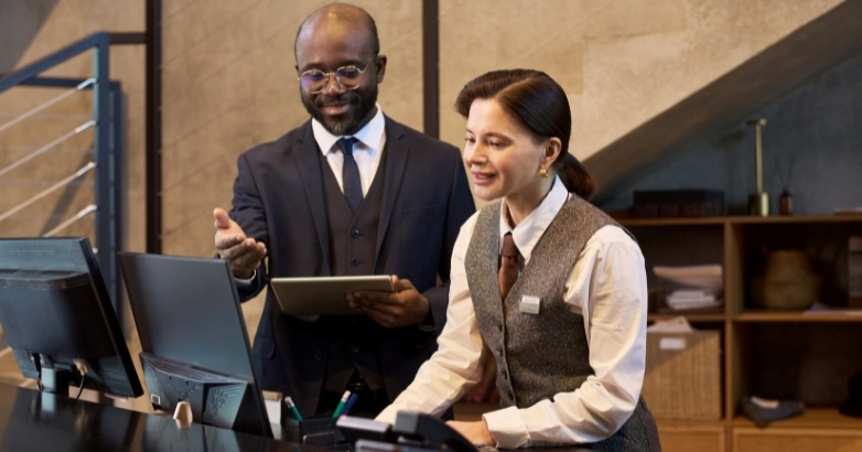 A man and woman are positioned in front of a desk, examining a tablet related to hotel reputation management software.